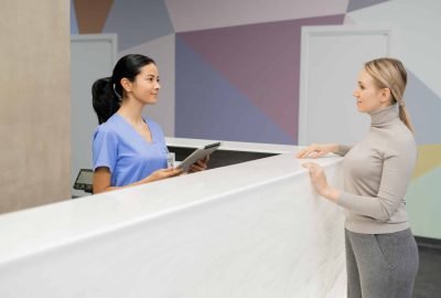 Young female assistant in blue uniform standing by reception counter and consulting patient in dental clinics lounge